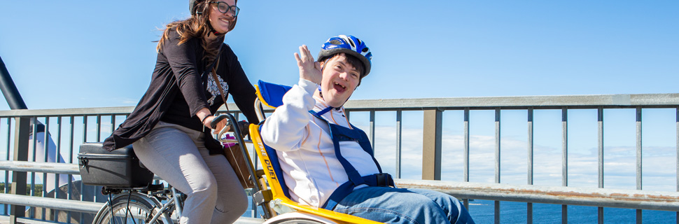 Ung man med downs syndrom sitter frampå en specialcykel som cyklas av en kvinna.