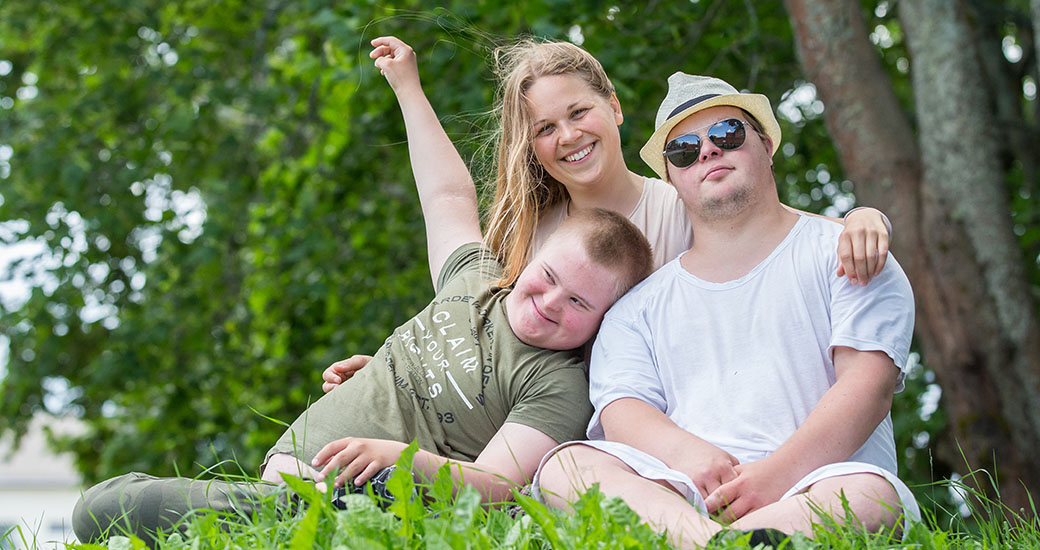 En ung kivnna och två unga killar med Downs syndrom sitter i gräset och håller om varandra.