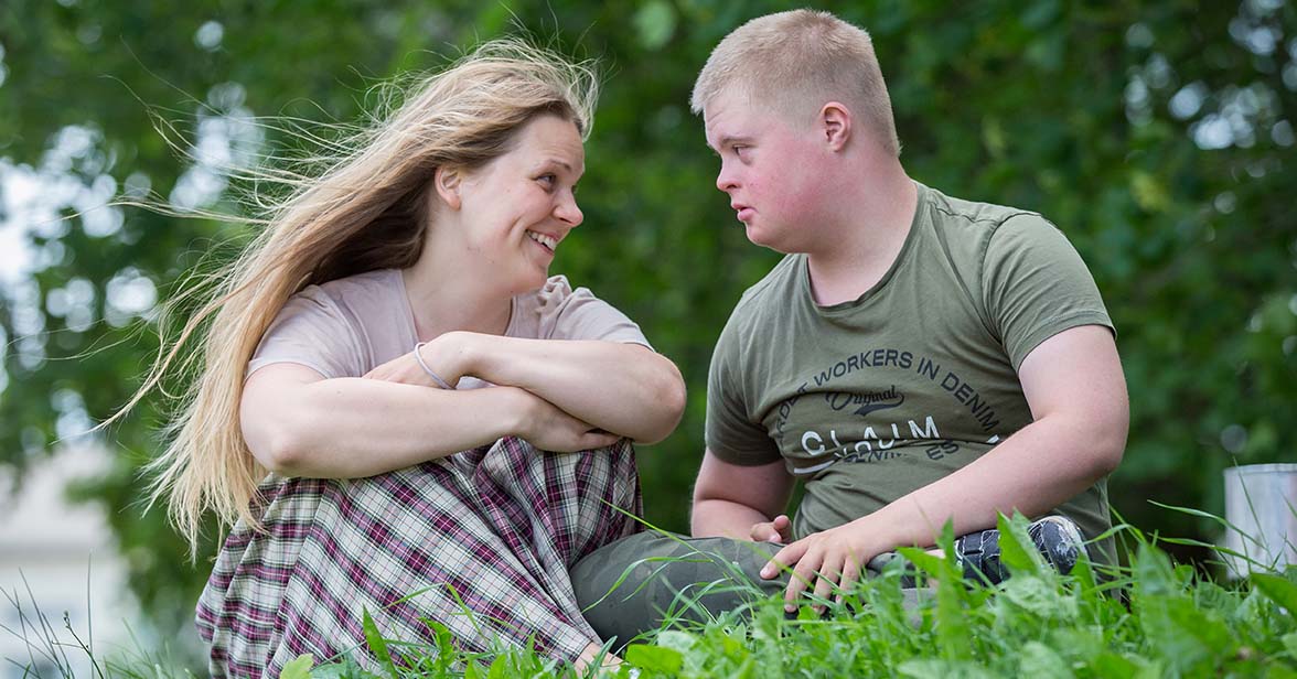 Två personer i sommargrönska sitter i gräset och tittar på varandra.