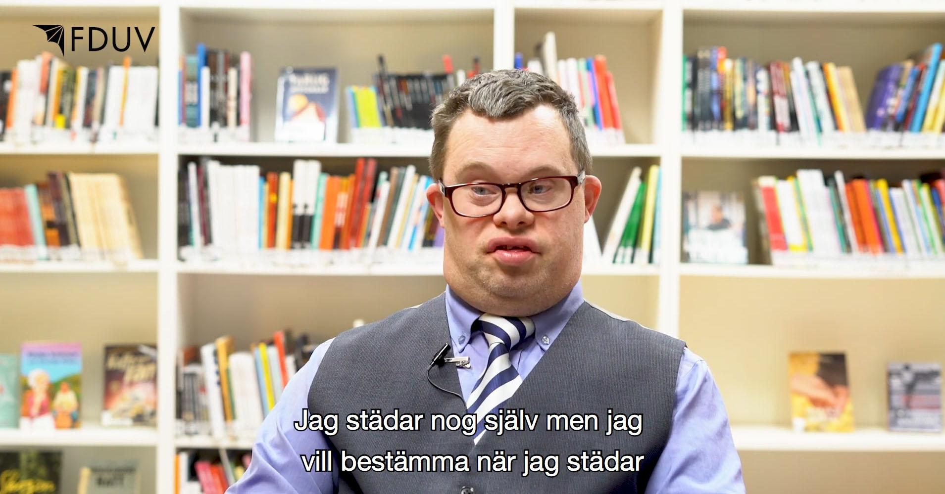 Skärmdump från film. Man med downs syndrom sitter framför en bokhylla. Text: Jag städar nog själv, men vill bestämma när jag städar