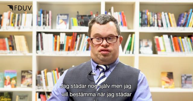 Skärmdump från film. Man med downs syndrom sitter framför en bokhylla. Text: Jag städar nog själv, men vill bestämma när jag städar