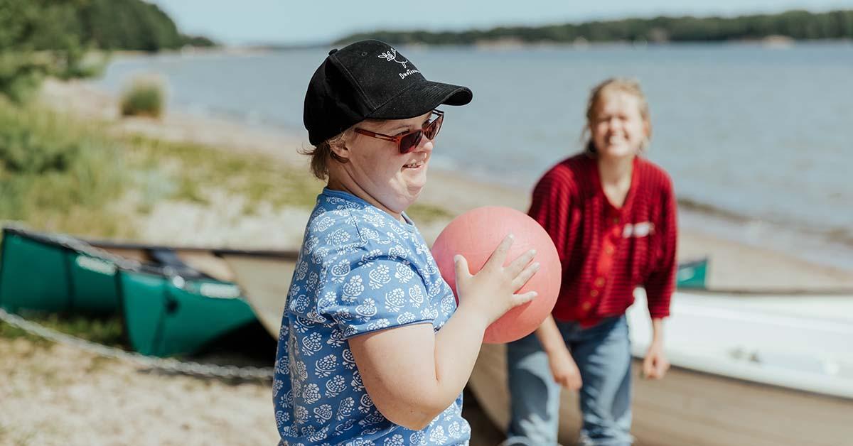 Person med downs syndrom leker på solig strand, i bakgrunden ledare.