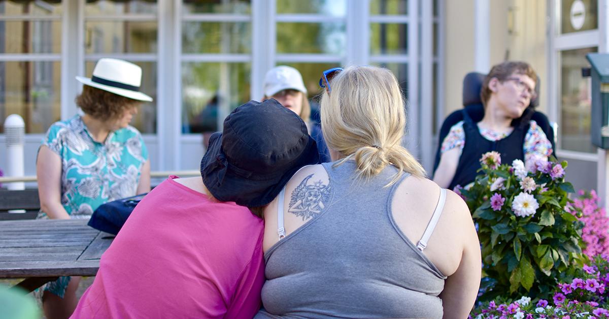 i förgrunden två personer som lutar mot varandra, i bakgrunden person i rullstol.