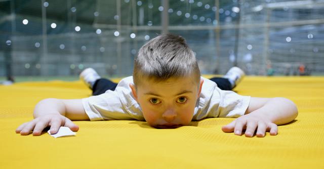 Kille med Downs syndrom ligger på mage och tittar rakt in i kameran.