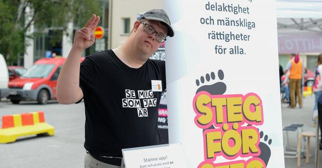 Person med Downssyndrom med t-shirt med texten Se mig som jag är vid Steg för Stegs rollup.