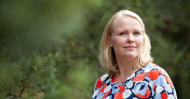 Anntte Tallberg-Haahtela med blommig blus mot grönskande bakgrund.