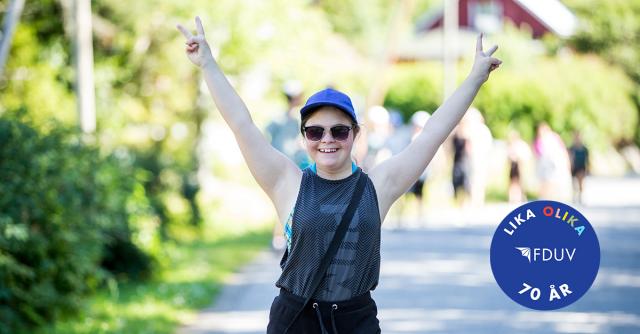 Ung kvinna med Down syndrom håller upp händerna och visar sägertecken i somrig miljö.